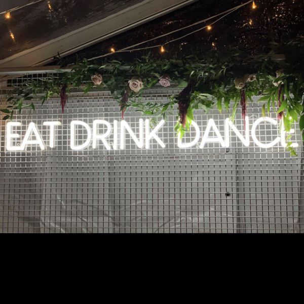 EAT DRINK DANCE large LED neon sign for events & weddings, shown on wire mesh with floral arrangement & fairy lights - photo from CustomNeon.co.uk