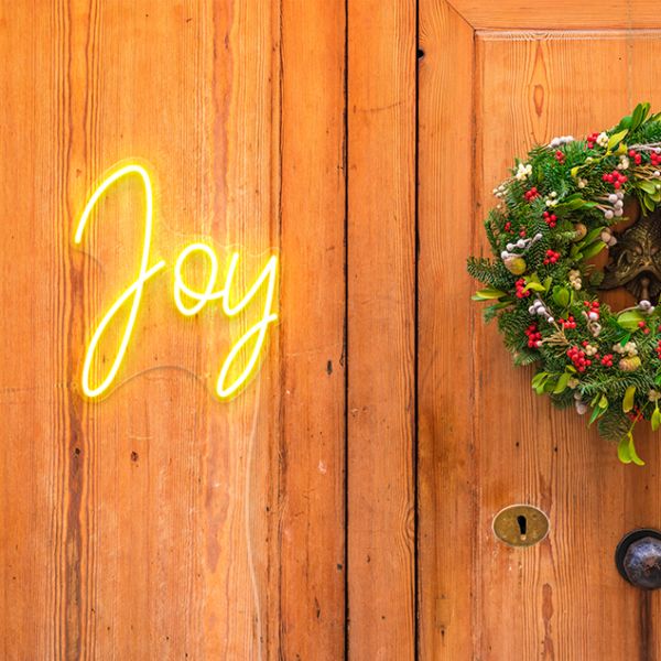 Joy light up sign by Custom Neon® in warm white shown hung near a Christmas wreath