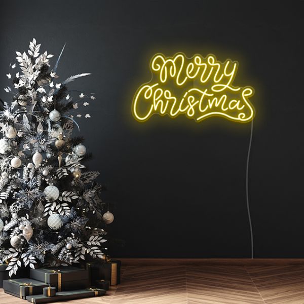 Yellow Merry Christmas sign in fancy script font against a dark wall next to a Christmas tree with monochrome decorations
