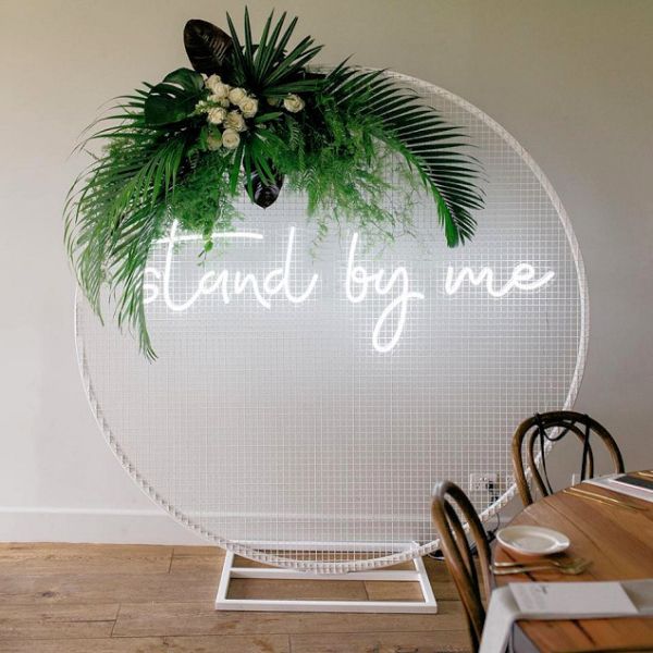 Stand by me LED neon wedding sign shown mounted on a mesh screen with a floral arrangement - photo from CustomNeon.co.uk
