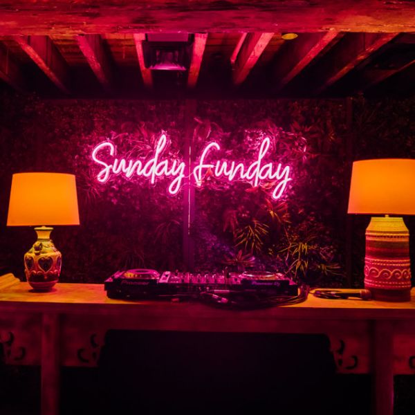 Sunday funday light up wall sign shown in pink displayed on a green wall in front of DJ decks - photo from Custom Neon by Neon Collective