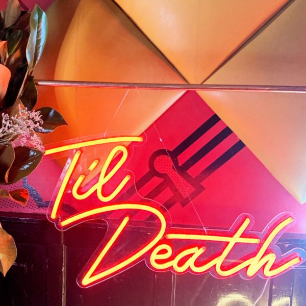 Til Death Neon Sign in LED flex shown in orange, hung from a frame at an event - photo CustomNeon.co.uk