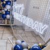 Custom Neon® Happy Birthday sign in white LED neon flex on an arched frame decorated with balloon garlands.