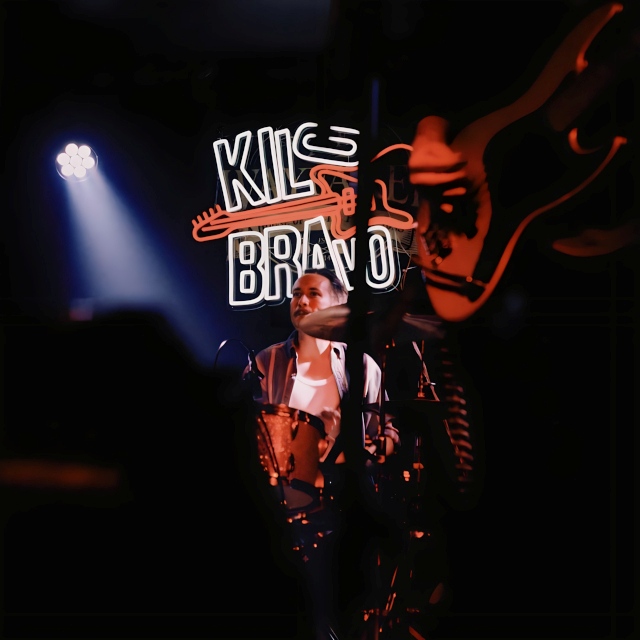 Drummer under stage lights in front of a Custom Neon® 'Kilo Bravo' band logo sign