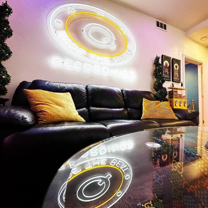 White and Yellow logo sign by Custom Neon® on a white wall above a black leather couch in the @qmadethebeat recording studio