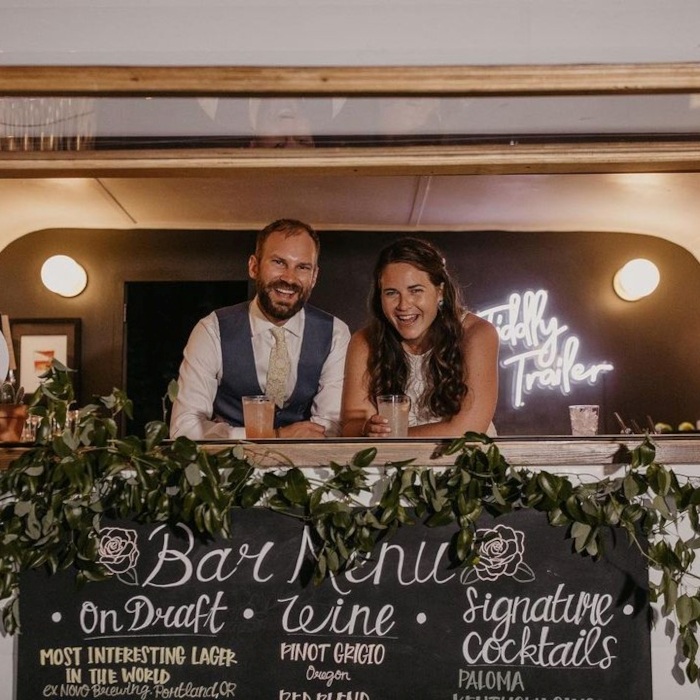 Bride and groom smiling from within the Tiddly Trailer bar van with a white Custom Neon® Tiddly Trailer sign in the background